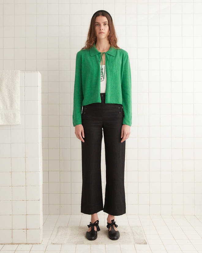 A person stands in a tiled room wearing a green Lodge Cardigan, white top, black pants, and black shoes.