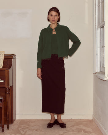 A woman stands indoors by a piano and window, wearing the Lodge Cardigan in dark green with a long black skirt for a cozy knitwear look.