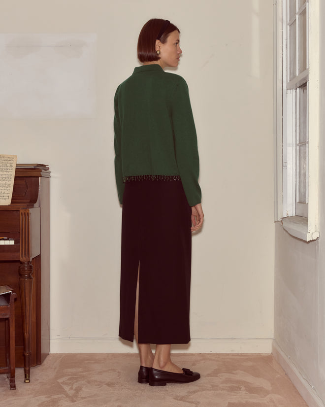 A woman in a Lodge Cardigan - Dark Green and black skirt stands indoors, facing away near a window and upright piano.