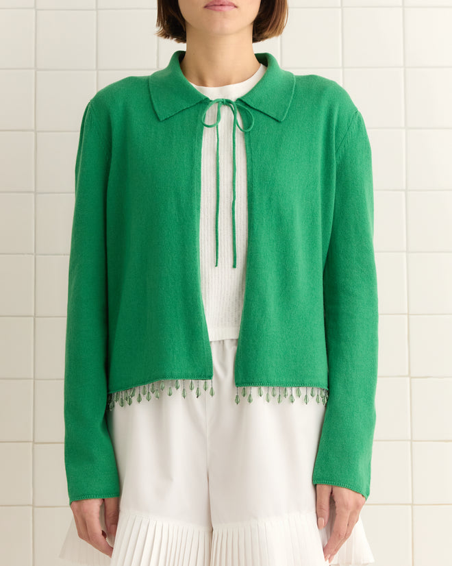 A person in a Lodge Cardigan - Green, featuring droplet beads, and a white pleated skirt stands gracefully against a tiled wall.