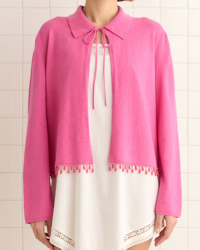 A person in a white dress and the Lodge Cardigan - Pink with a tie collar and droplet bead details stands gracefully against a tiled wall.