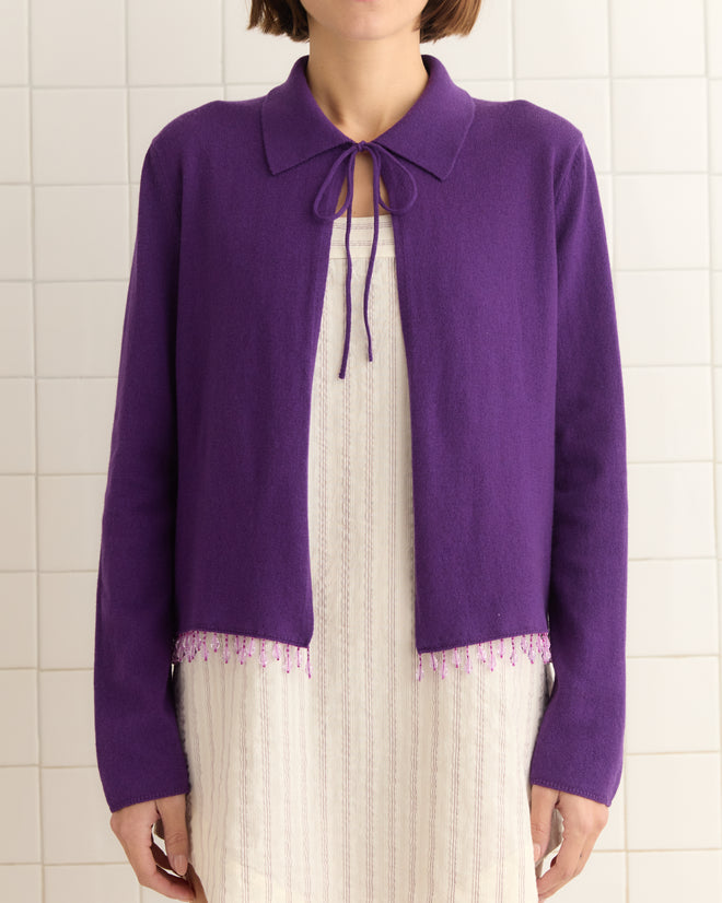 A person dressed in a Lodge Cardigan - Plum and a white dress stands elegantly against a tiled wall.