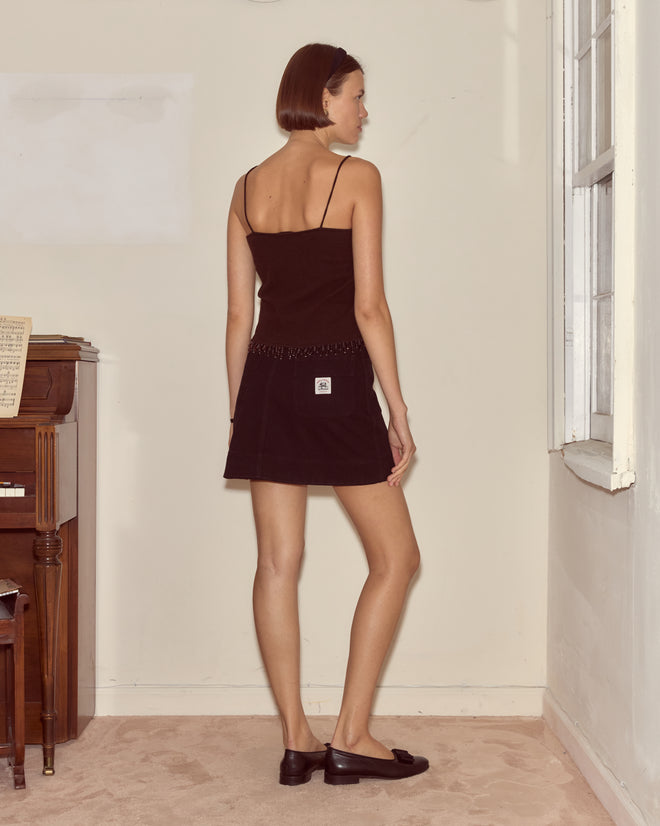 Woman in a dark brown Lodge Tank and skirt stands by a white wall, facing away, next to a piano and window.