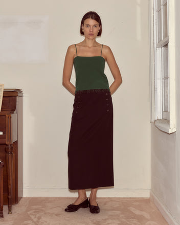 A woman stands indoors by a window wearing the Lodge Tank in Dark Green, styled with a black maxi skirt and black flats.