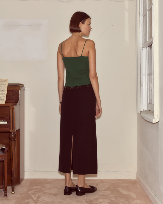 A woman with short hair stands indoors, facing away, wearing the Lodge Tank in dark green and black pants near a piano and window.