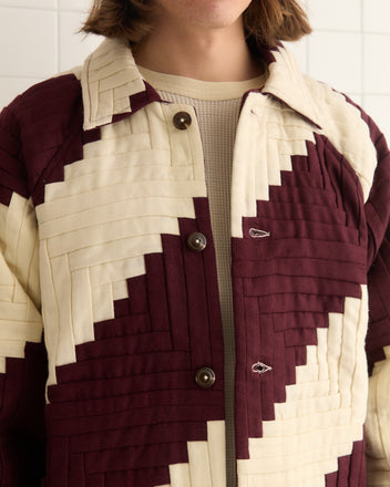 A person in a Log Cabin Suiting Coat with a maroon and cream geometric quilted pattern and embroidered logo stands against a tiled wall.