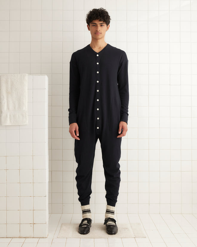 A person wearing a navy jumpsuit made of organic cotton and Long Johns - Black stands in a tiled room with a towel hanging on the wall.