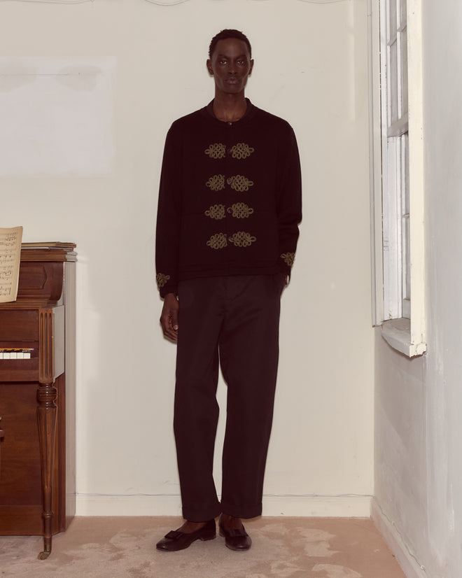 A man in dark clothing wears the Looped Frog Cardigan, standing indoors by a piano and window, facing forward with a neutral expression.