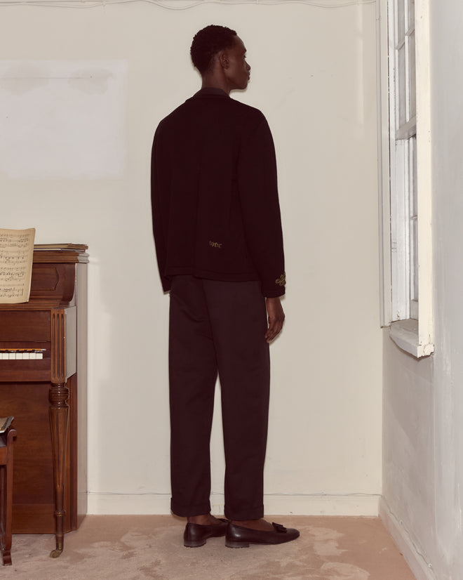 A person wearing the Looped Frog Cardigan stands facing the corner of a room near a piano and a window.