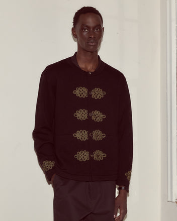 A person wearing the Looped Frog Cardigan, a black knitwear jacket featuring gold embroidery and trompe l'oeil frog closures, stands against a light-colored wall.