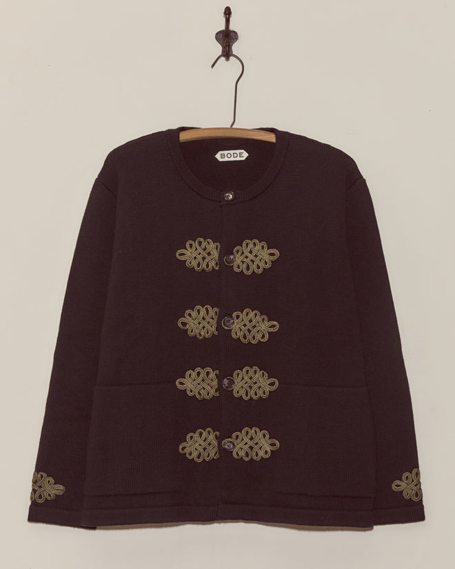 A black BODE Looped Frog Cardigan featuring merino wool, decorative gold embroidery, and trompe l'oeil frog closures hangs on a wooden hanger against a plain wall.