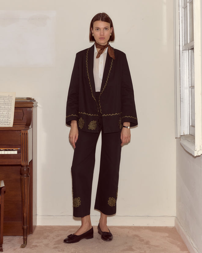 A woman stands indoors by a piano, wearing the Looped Frog Jacket—a black shawl-collar style adorned with bronze bullion—and a brown neck scarf.