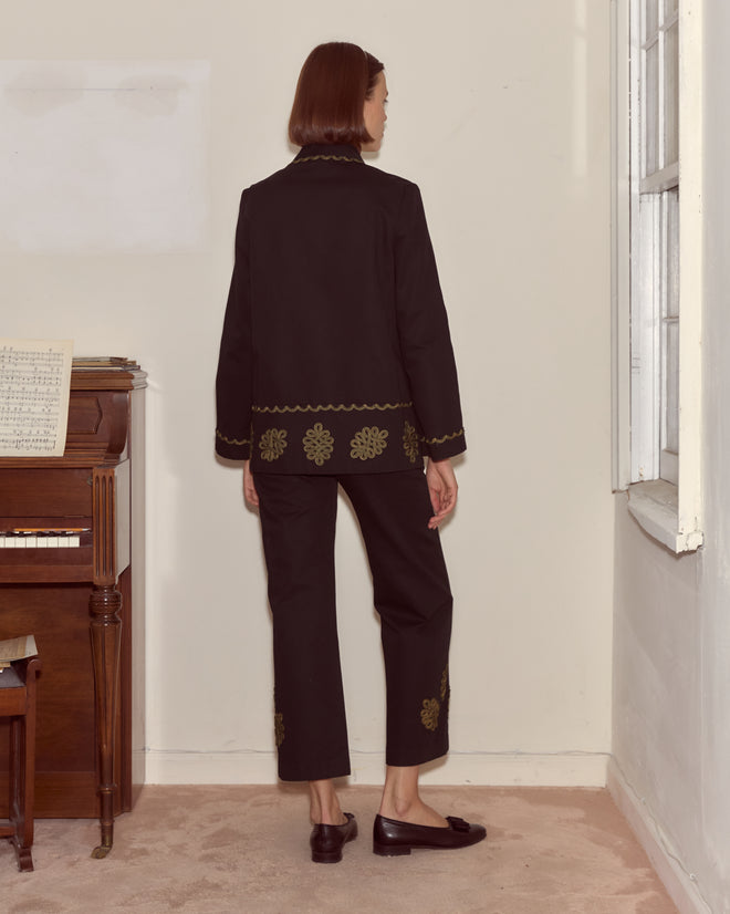 A woman in the Looped Frog Jacket, featuring a black shawl collar and gold embroidery, stands facing away beside a piano in a softly lit room.