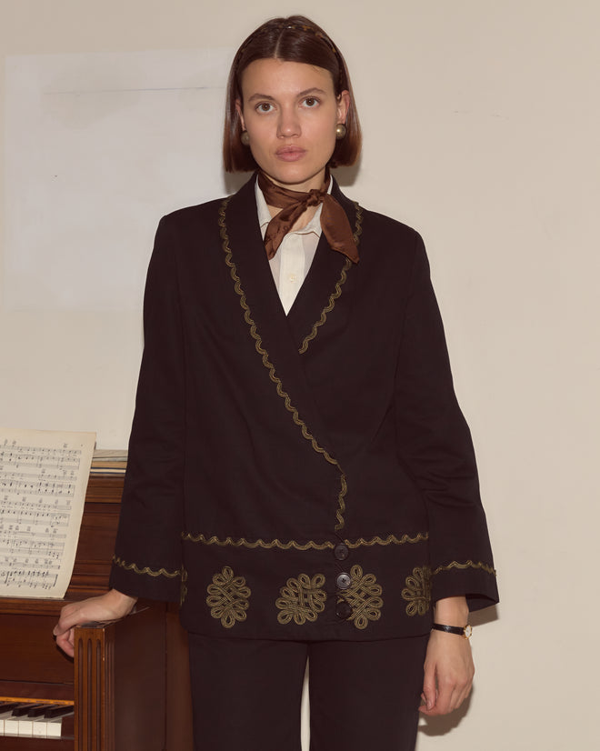 A woman in the Looped Frog Jacket, featuring a black shawl collar and bronze bullion details, stands by a piano with sheet music, paired with a brown scarf and white shirt.