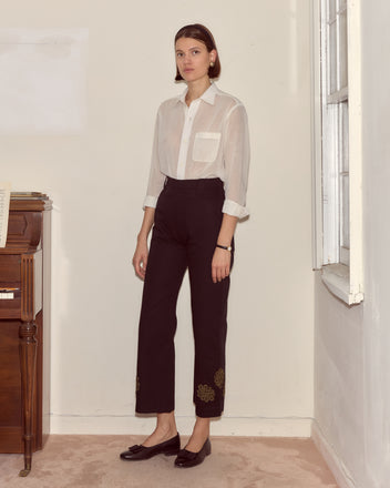 Indoors by a piano, a woman wears a white shirt with smile pockets, black Looped Frog Trousers in cotton twill, and black shoes.