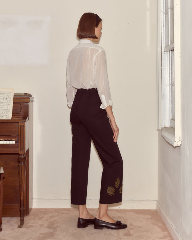A woman stands indoors facing away, wearing a sheer white shirt and Looped Frog Trousers beside a piano and window.