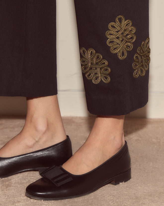 Person wearing Looped Frog Trousers—black cotton twill with gold embroidery at the hem and smile pockets—paired with black low-heeled shoes featuring a bow.