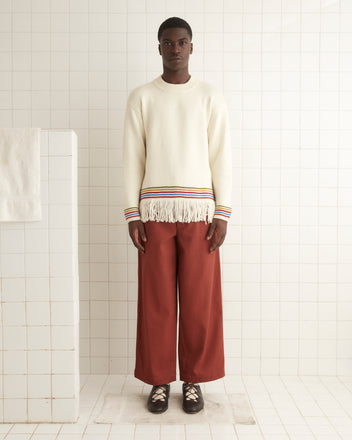 A man stands in a tiled room wearing the Loop Edge Pullover, a cream merino wool sweater with fringe and colorful stripes at the cuffs and hem, paired with wide rust-colored pants and dark shoes.