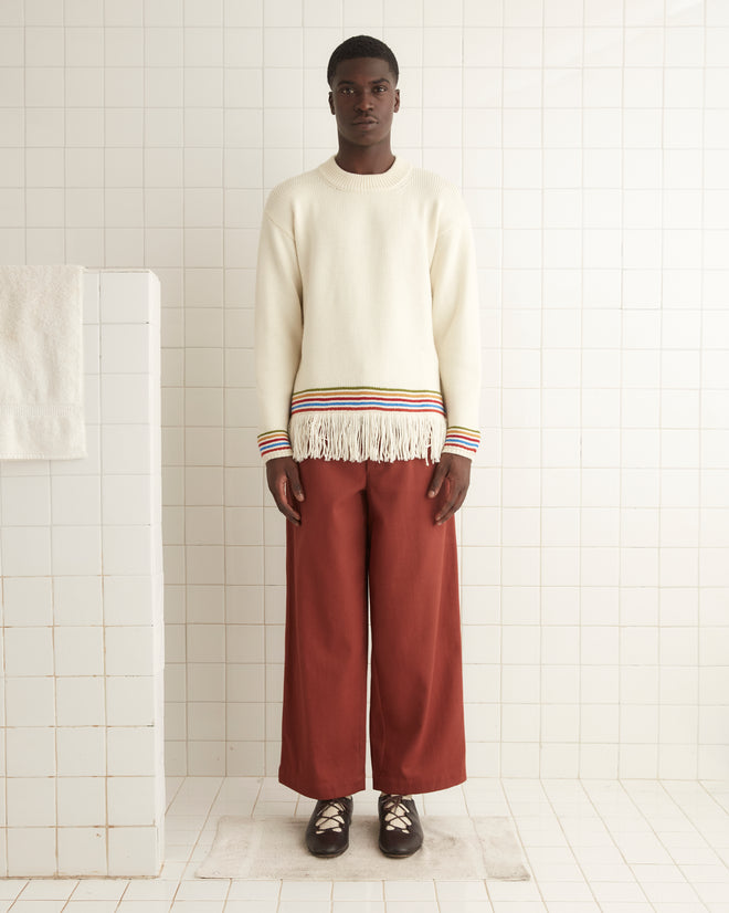 A man stands in a tiled room wearing the Loop Edge Pullover, a cream merino wool sweater with fringe and colorful stripes at the cuffs and hem, paired with wide rust-colored pants and dark shoes.