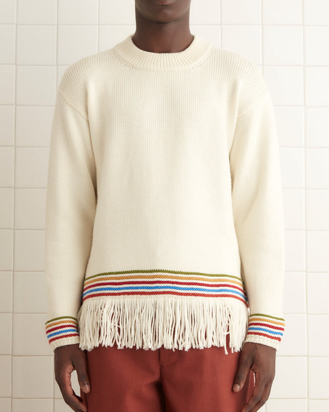 A man stands in a tiled room wearing the Loop Edge Pullover, a cream merino wool sweater with fringe and colorful stripes at the cuffs and hem, paired with wide rust-colored pants and dark shoes.