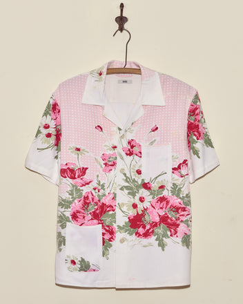 The Lovely Garden Short Sleeve Shirt (XS/S), featuring a pink and red floral print, is displayed on a wooden hanger against a beige wall.