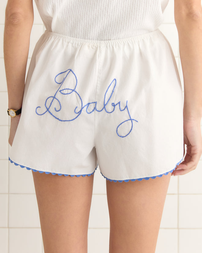 A person is against a tiled wall wearing Lucky Baby Shorts, featuring "Baby" in blue script and a scalloped hem, resembling delicate baby bloomers.