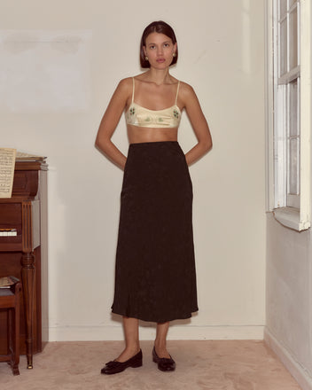 Indoors by a piano, a woman wears the Lucky Draw Bralette—hand-embroidered silk—paired with a long dark skirt and black shoes subtly featuring a playing card motif, gazing at the camera.