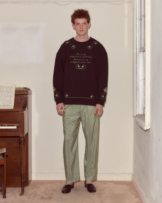 A man in the Lucky Draw Pullover, featuring a subtle hand-embroidered card suit motif, and green striped pants stands by a piano and window.