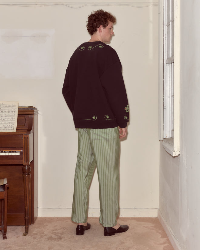 A man wearing the Lucky Draw Pullover, a hand-embroidered sweatshirt with a card suit motif, and green striped pants stands by a window in a sparse room with a piano, facing away.