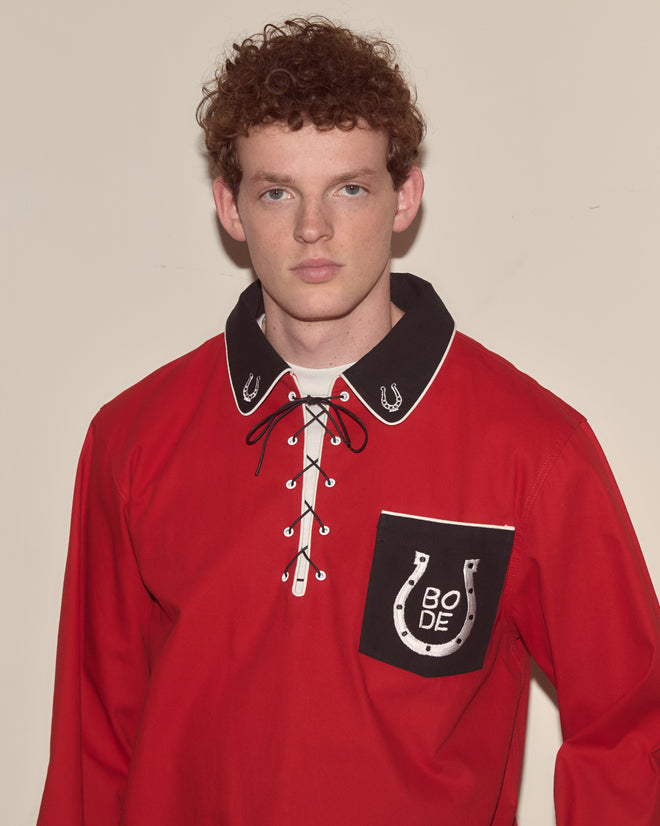 A young man with curly hair wears the Lucky Horseshoe Pullover, a red Western shirt featuring a lace-up collar and embroidered horseshoes on the pocket.