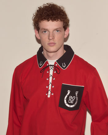 A young man with curly hair wears the Lucky Horseshoe Pullover, a red Western shirt featuring a lace-up collar and embroidered horseshoes on the pocket.