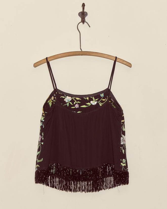 The Luna Rose Top - S is a unique black camisole featuring floral embroidery and beaded fringe, styled like an embroidered shawl, displayed on a wooden hanger against a beige wall.
