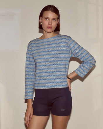 Woman wears the Lyric Long Sleeve in blue jacquard stripes with black shorts, standing against a light-colored wall.