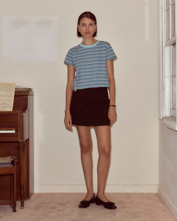 A woman in the Lyric Cropped Tee and a black skirt stands indoors by a piano and window, creating the ambiance of an Expressionist musical.
