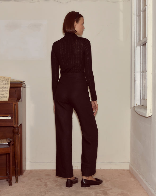 A woman in black pants and a Louise Pullover - Black stands facing a wall near a piano and a window.