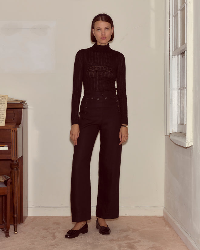 A woman in a black Louise Pullover and high-waisted pants stands indoors by a piano and a window.
