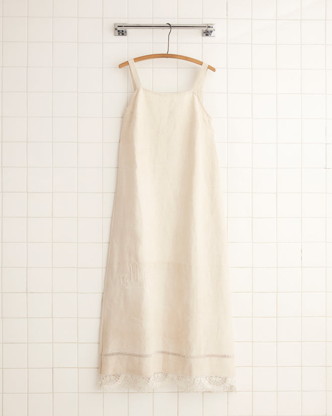 M.C. Monogram Dress in beige, featuring a crochet lace hem, displayed on a wooden hanger against a tiled wall, evoking the charm of an antique French bedspread.