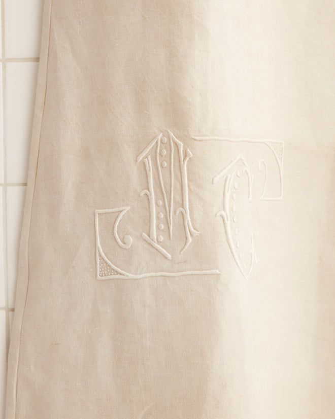 M.C. Monogram Dress in beige, featuring a crochet lace hem, displayed on a wooden hanger against a tiled wall, evoking the charm of an antique French bedspread.