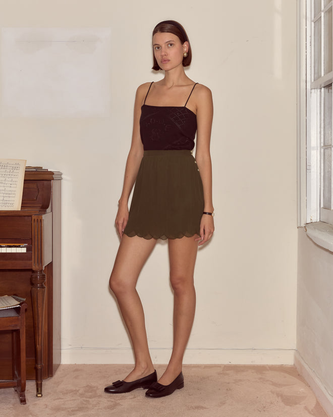 A woman stands indoors by a piano, wearing a black top, black flats, and the Mab Skirt—a crinkled georgette olive skirt.