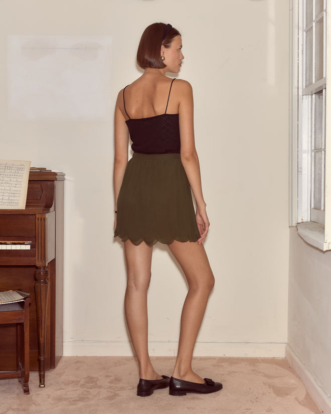 A woman wearing the Mab Skirt in olive silk and a black top stands indoors, facing away by a piano and window.