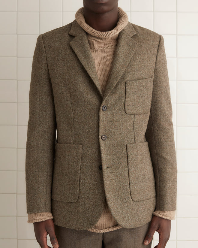 Against a wooden background, a person is wearing the Macnamara Suit Jacket - 40 in brown over a turtleneck, evoking classic 1960s suiting fabric with its 100% wool composition.