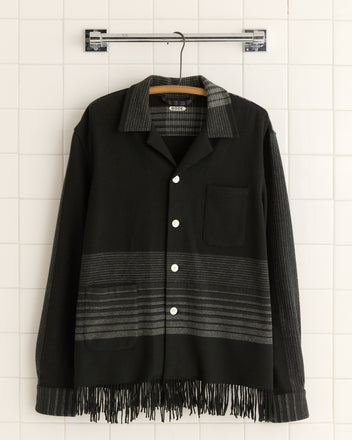 The Magnetite Fringe Overshirt - S/M in black, crafted from 100% wool, is displayed on a hanger against a tiled wall.