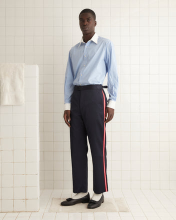 A person in a light blue shirt and Major League Suiting Trousers with a satin handfeel, featuring a red stripe, stands in a tiled room.