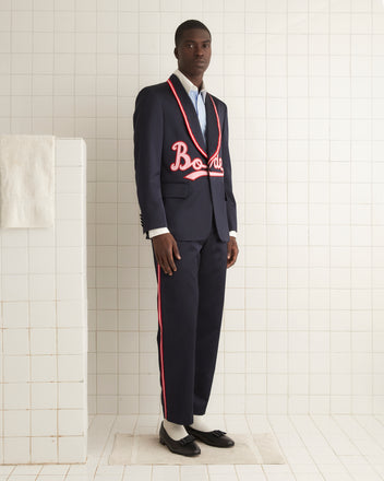 A person wearing the Major League Suit Jacket, a stylish dark suit with red trim and embroidered text, featuring a classic shawl collar, stands against a brown curtain backdrop.