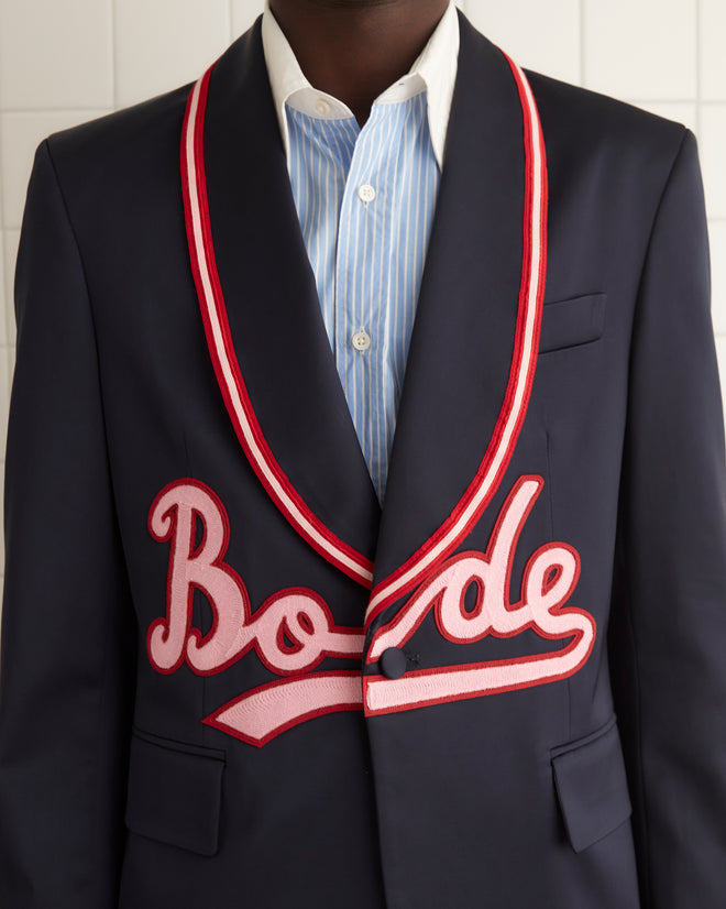 A person wearing the Major League Suit Jacket, a stylish dark suit with red trim and embroidered text, featuring a classic shawl collar, stands against a brown curtain backdrop.