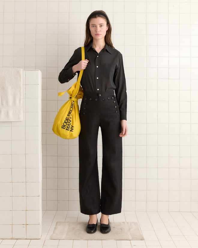 A person in a black outfit with a Manhattan Island Bag, featuring a yellow waxed cotton finish and adjustable cinch tie, stands against a white tiled wall.