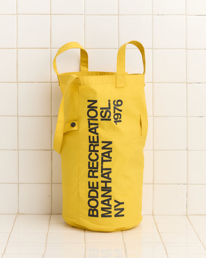 The Manhattan Island Bag is a yellow cylindrical seabag made from waxed cotton, featuring the text "BODE RECREATION MANHATTAN ISL. 1976" and displayed against a tiled wall background. It's a stylish and functional souvenir with an adjustable cinch tie for convenience.