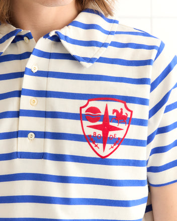 A person wears the Margate Polo, a blue and white striped cotton polo with a red embroidered crest on the chest, evoking the classic French Navy style.