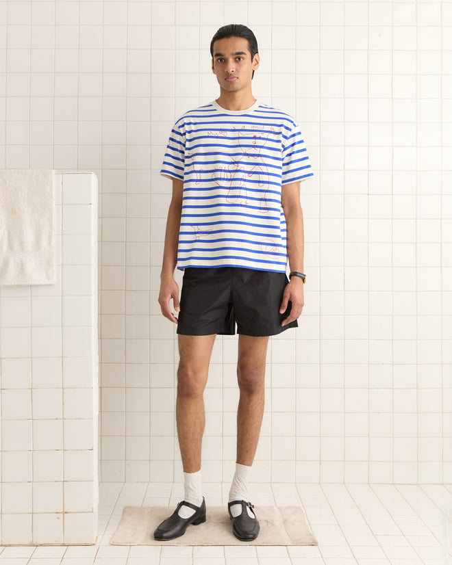 A person wearing the Margate Tee and black shorts stands in a white tiled room, hands resting calmly at their sides.