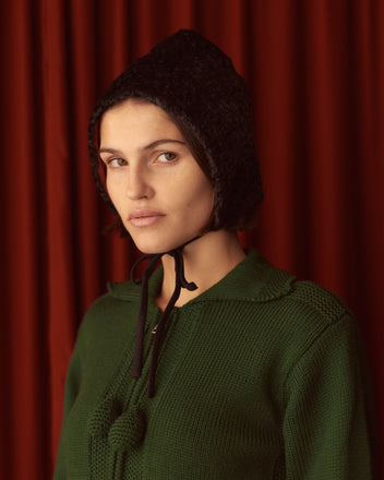 A person in a green knit outfit, enhanced by chic accessories like the Faux Fur Tie Cap in black, poses against a red curtain backdrop.